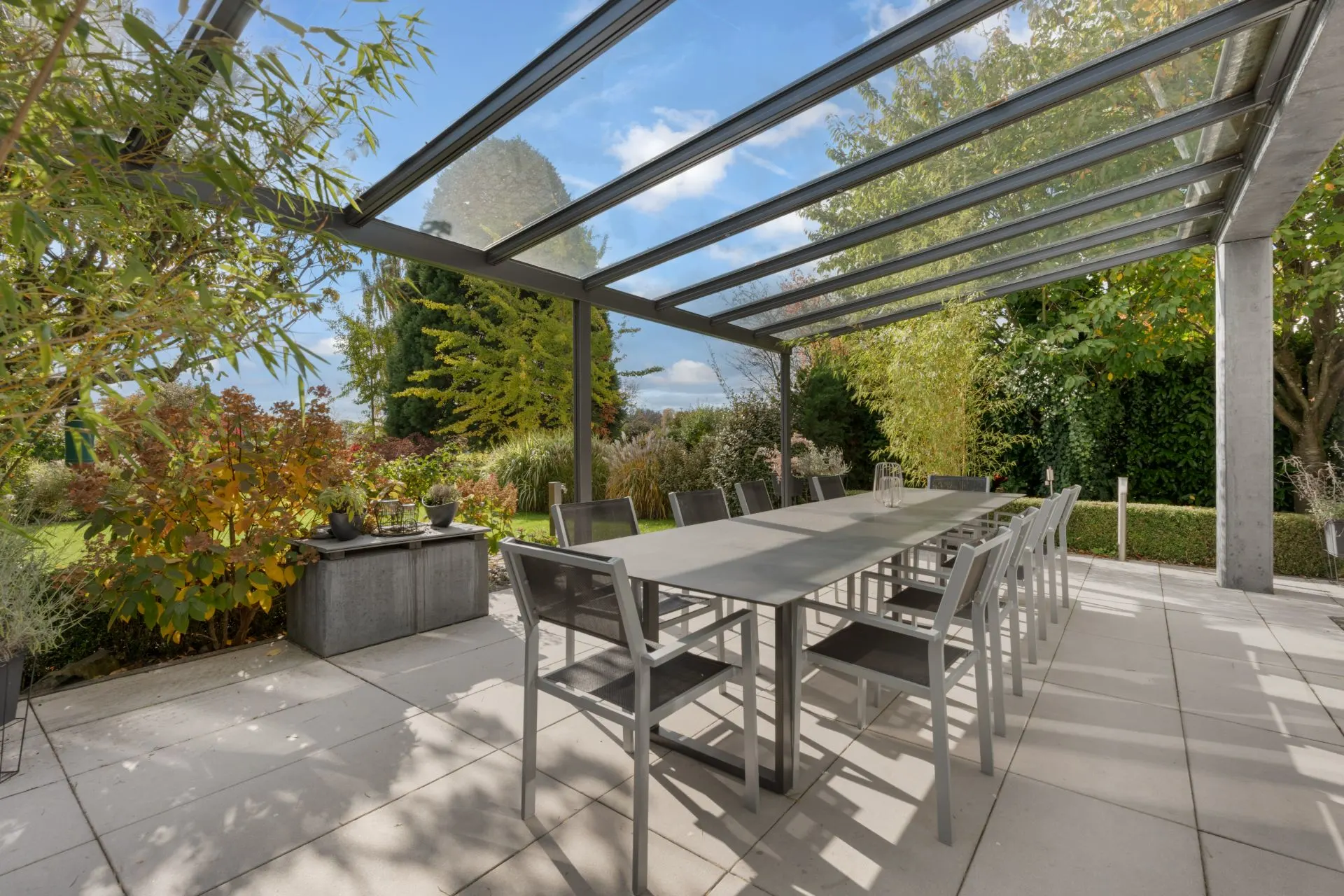 Überdachte Terrasse mit großem Tisch und Stühlen, umgeben von Gartenpflanzen und Bäumen.