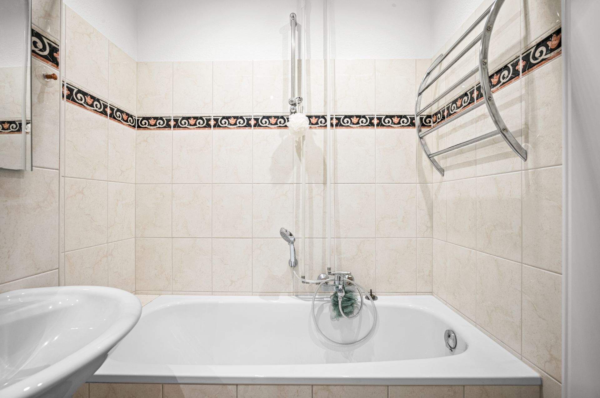 Badezimmer mit Badewanne, Duschstange, Handtuchhalter und hellen Fliesen mit Zierleiste.