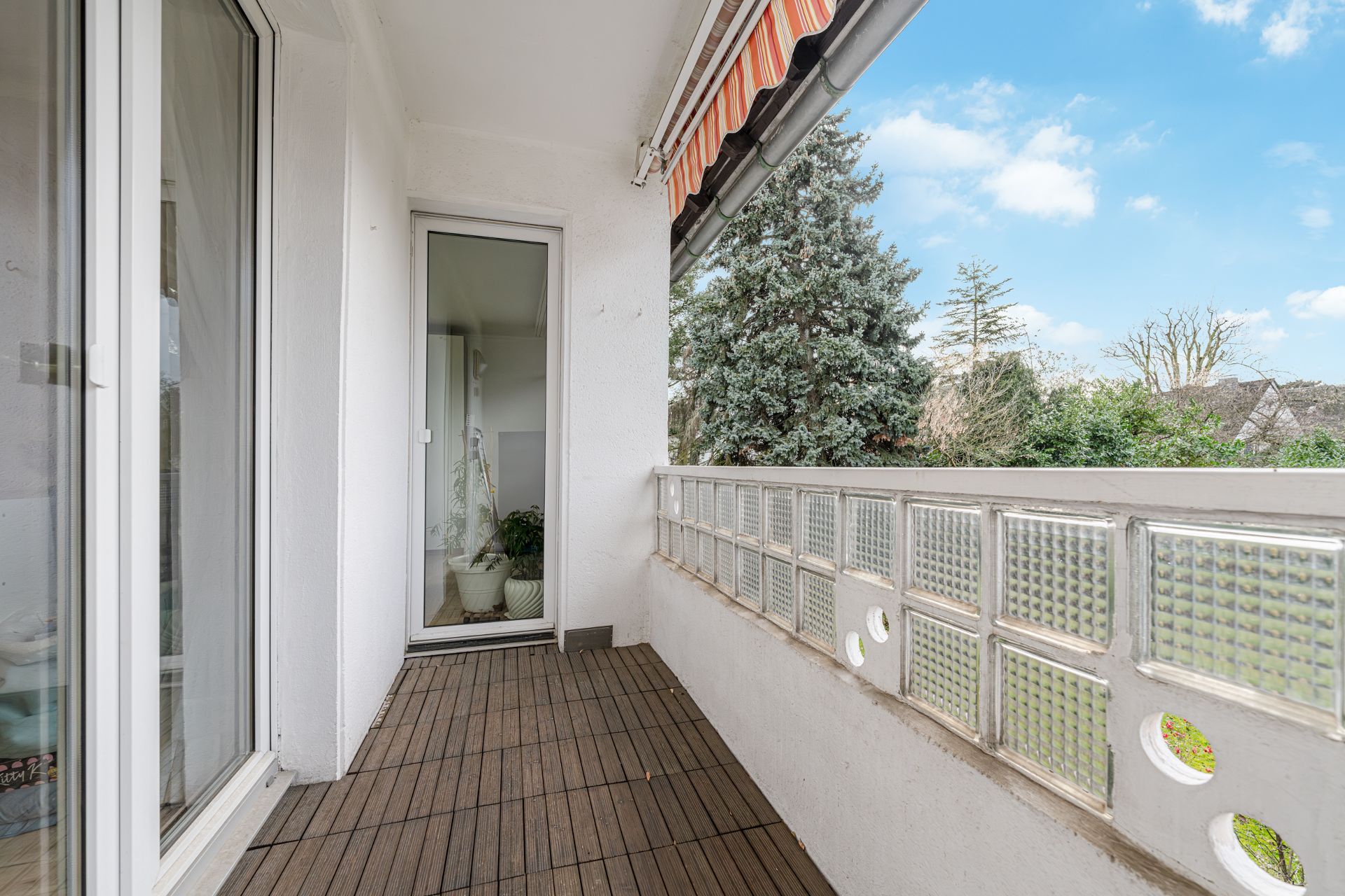 Balkon mit Holzboden, Glasbaustein-Geländer und Markise, Blick auf Bäume.