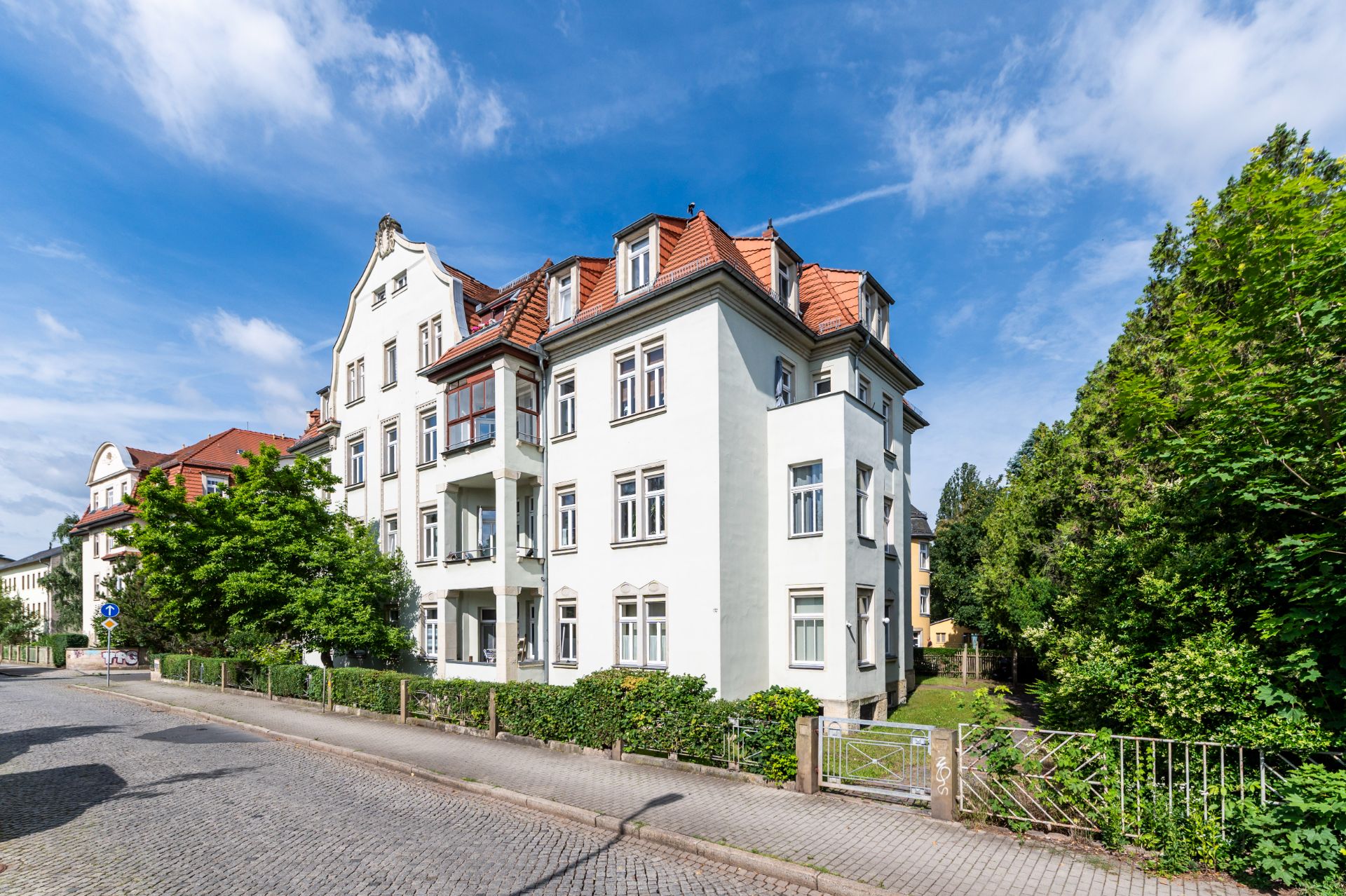 Mehrstöckiges Wohnhaus mit hellen Fassaden und rotem Dach an einer ruhigen Straße, umgeben von Bäumen.