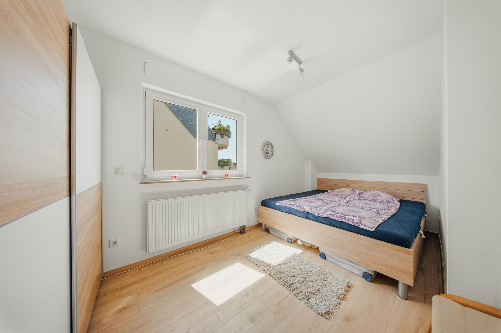 Helles Schlafzimmer mit Holzboden, Doppelbett, Fenster und Wanduhr.