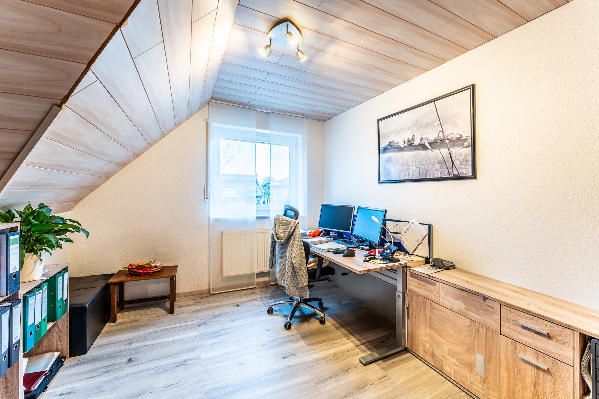 Helles Arbeitszimmer mit Schreibtisch, Monitoren, Regal und schräger Holzdecke.