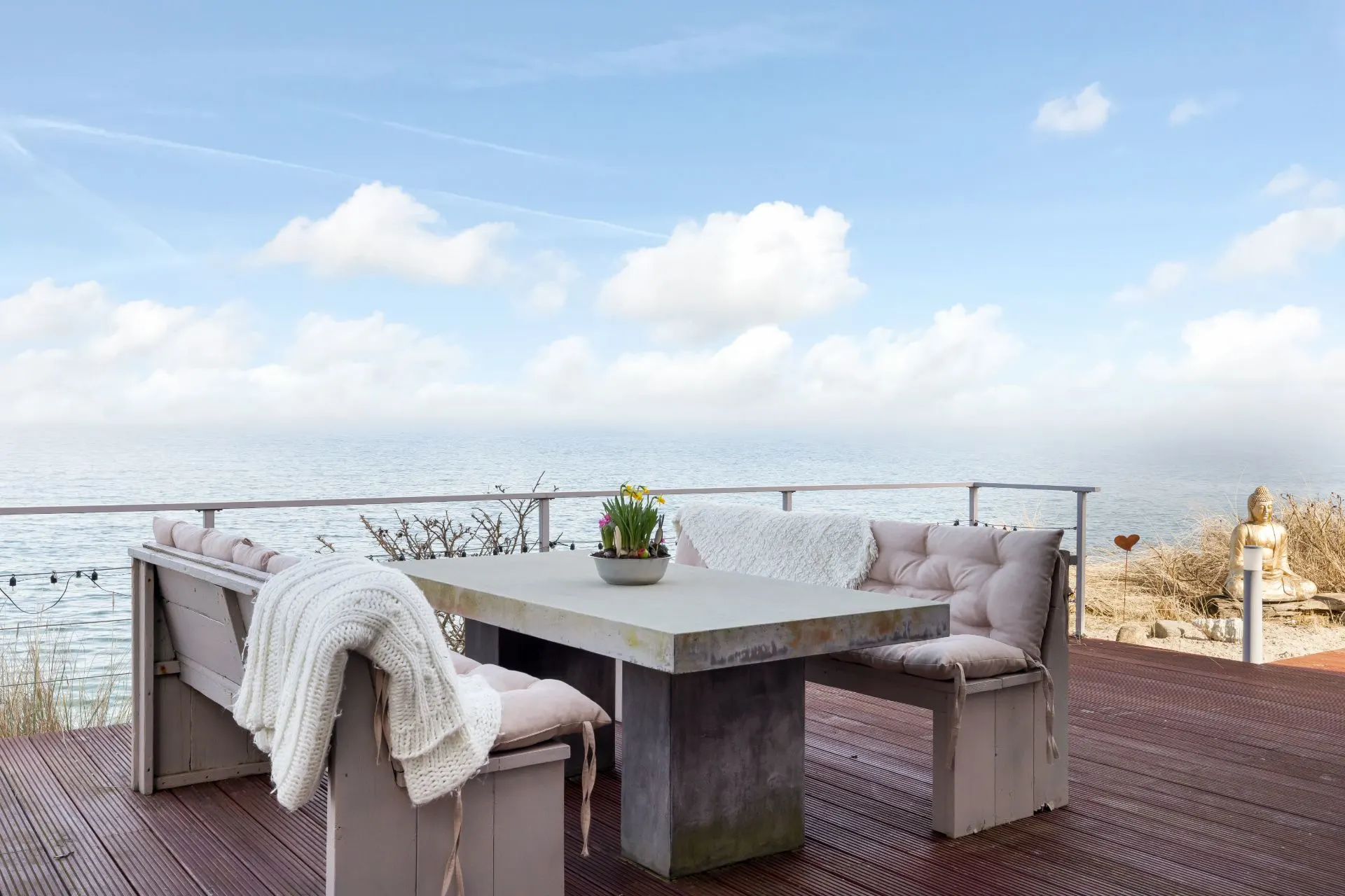 Terrasse mit Sitzbank, Tisch, Kissen und Blick auf das Meer bei klarem Himmel.