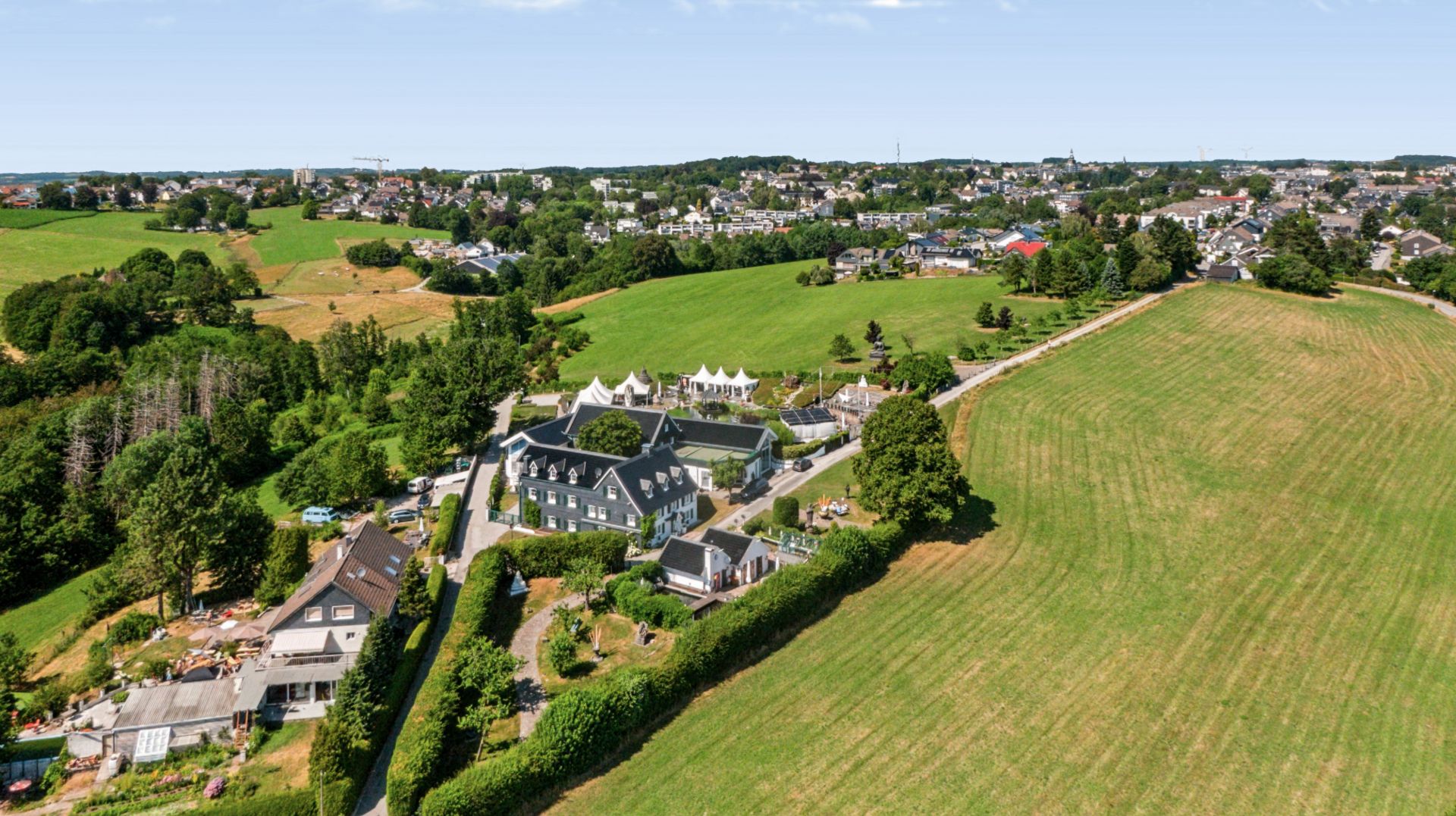 Luftaufnahme mehrerer Häuser am Rand eines großen grünen Feldes mit Bäumen und weitem Blick.