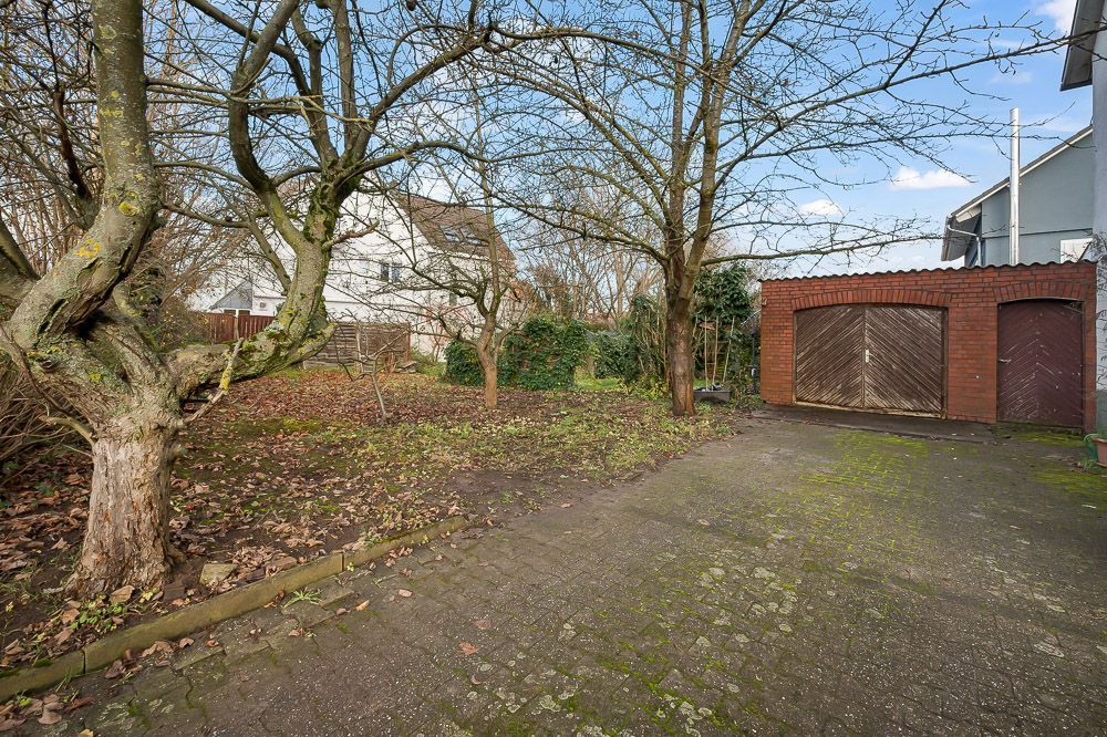 Gepflasterter Hof mit zwei Bäumen, einer Garage aus rotem Backstein und angrenzendem Gartenbereich.