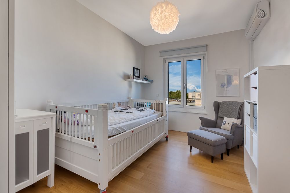 Helles Kinderzimmer mit Gitterbett, Sessel und Fenster, Holzboden und weißer Einrichtung.