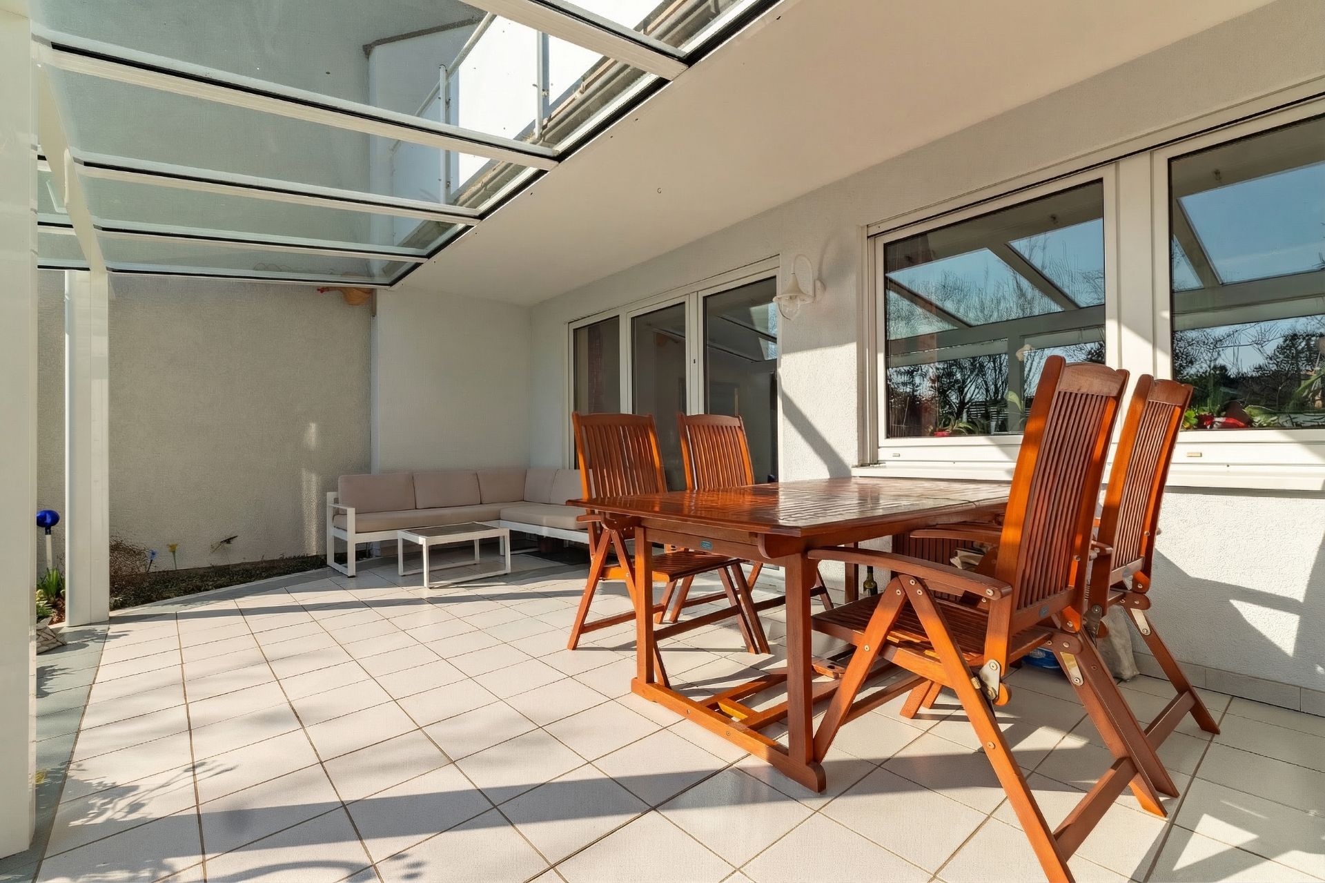 Überdachte Terrasse mit Holztisch, vier Stühlen und einer Sitzgruppe auf gefliestem Boden.