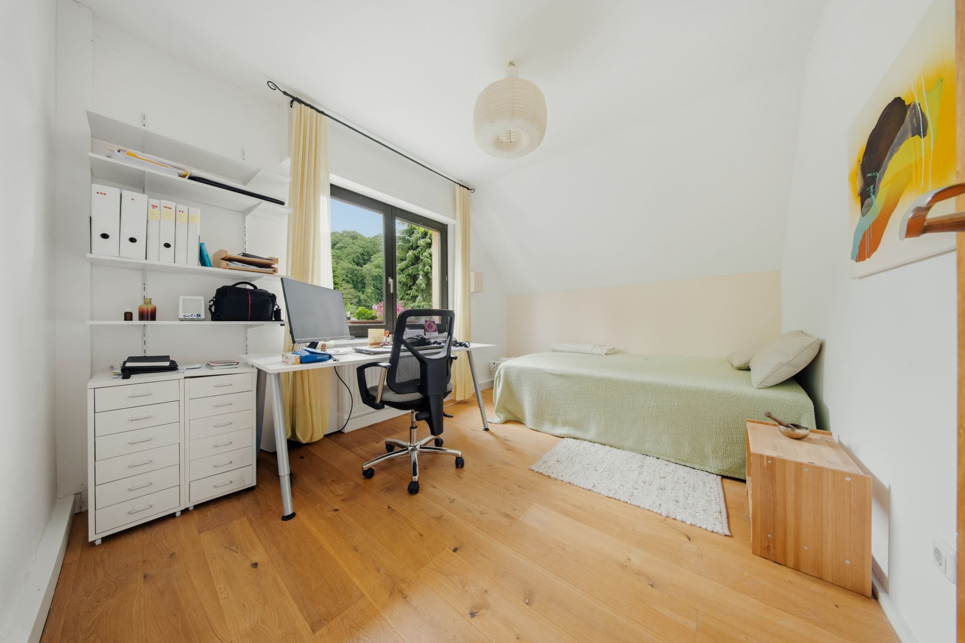 Helles Zimmer mit Schreibtisch, Stuhl, Bett und großem Fenster, Holzboden und Regal.