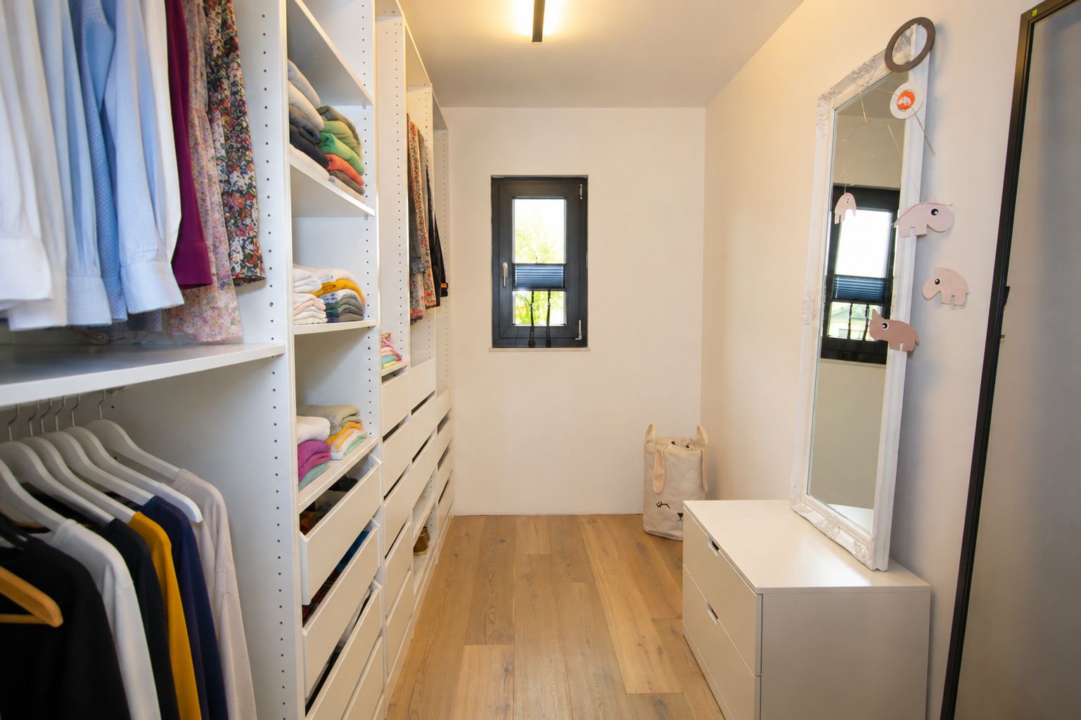 Begehbarer Kleiderschrank mit Regalen, Kleidung, Kommode und Spiegel, Holzfußboden, Fenster am Ende.