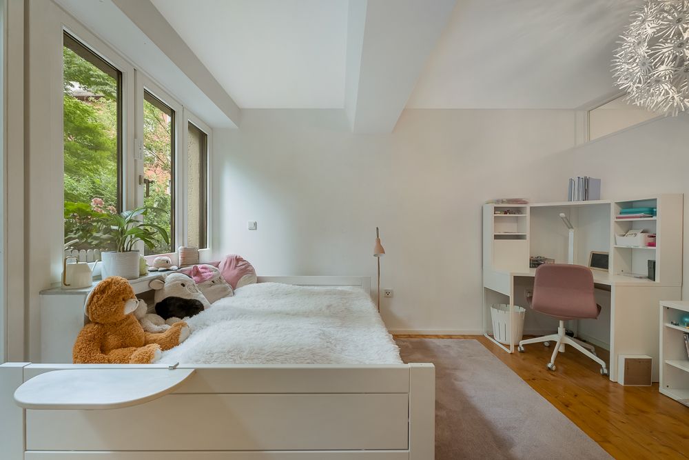 Helles Schlafzimmer mit Einzelbett, Kuscheltieren, großem Fenster und Schreibtisch mit Stuhl.