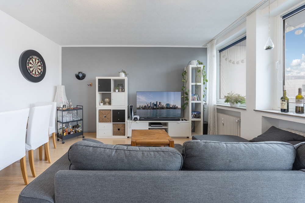 Wohnzimmer mit grauem Sofa, Fernseher, Regal, Esstisch, Dartscheibe und großen Fenstern.