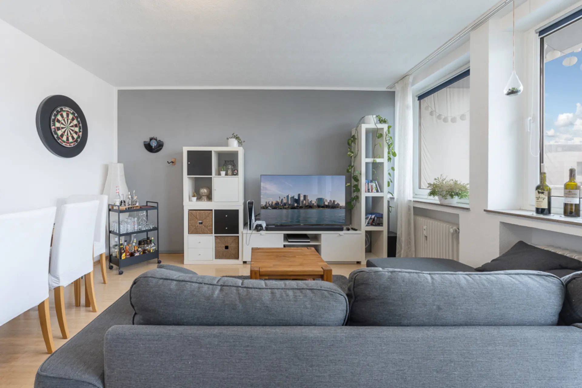 Wohnzimmer mit grauem Sofa, Fernseher, Regal, Esstisch, Dartscheibe und großen Fenstern.