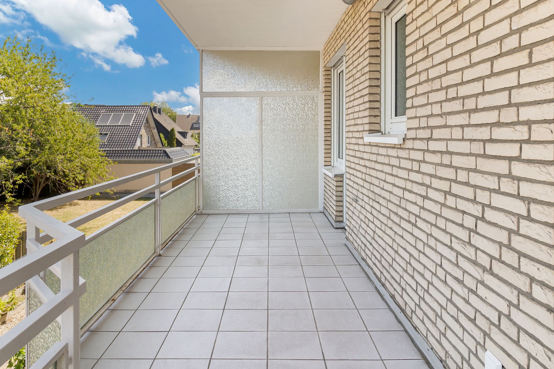 Großer Balkon mit Fliesenboden, Geländer und Ziegelwand, Blick auf Nachbarhäuser und Bäume.