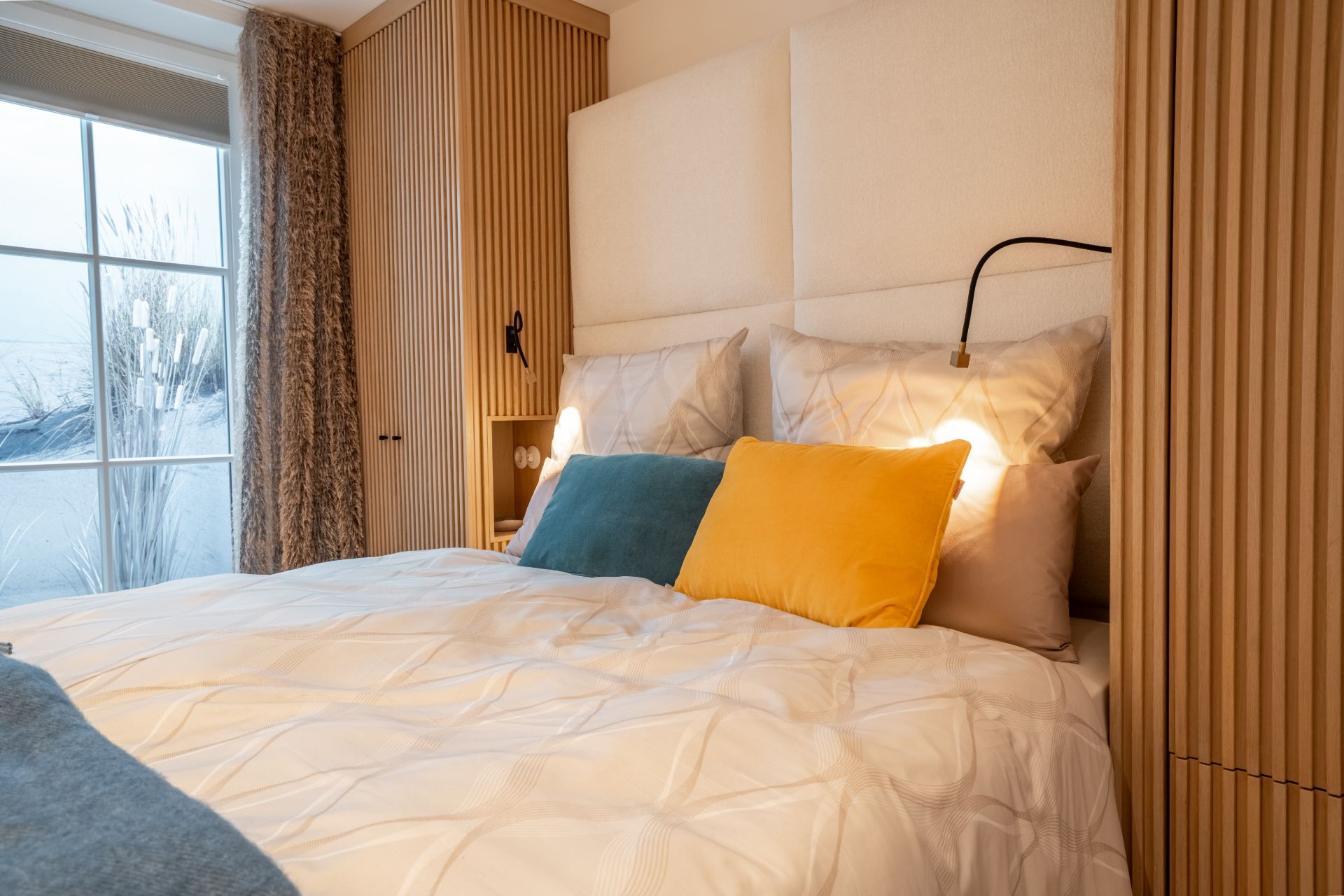 Modernes Schlafzimmer mit Doppelbett, bunten Kissen und großem Fenster mit Vorhang.