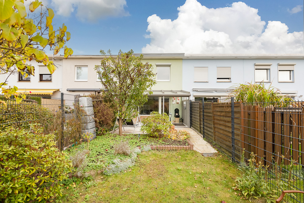 Reihenhaus mit kleinem Garten, Baum, Zaun und Terrasse im Hinterhof bei Tageslicht.