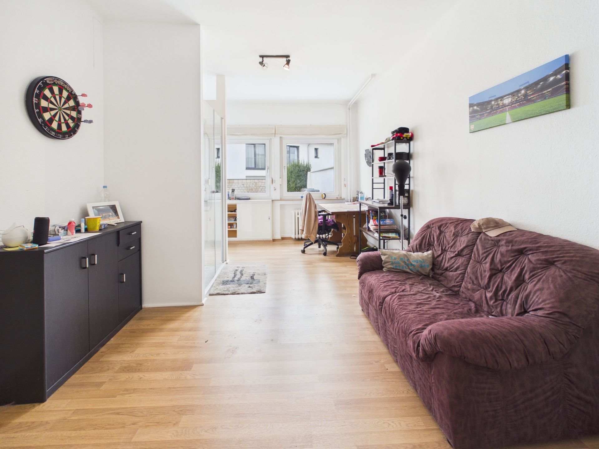 Helles Wohnzimmer mit braunem Sofa, Schrank, Dartscheibe und Schreibtisch am Fenster.