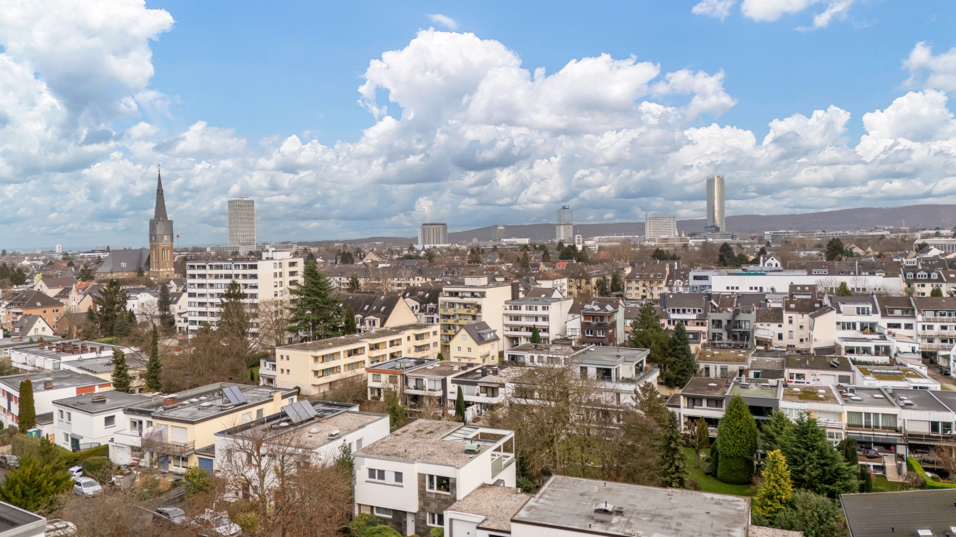 Blick über eine Wohngegend mit Häusern, Bäumen und einem Kirchturm unter blauem Himmel.
