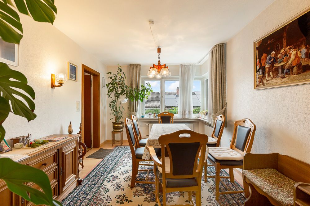 Esszimmer mit Holztisch, sechs Stühlen, Teppich, Pflanzen und großem Fenster.