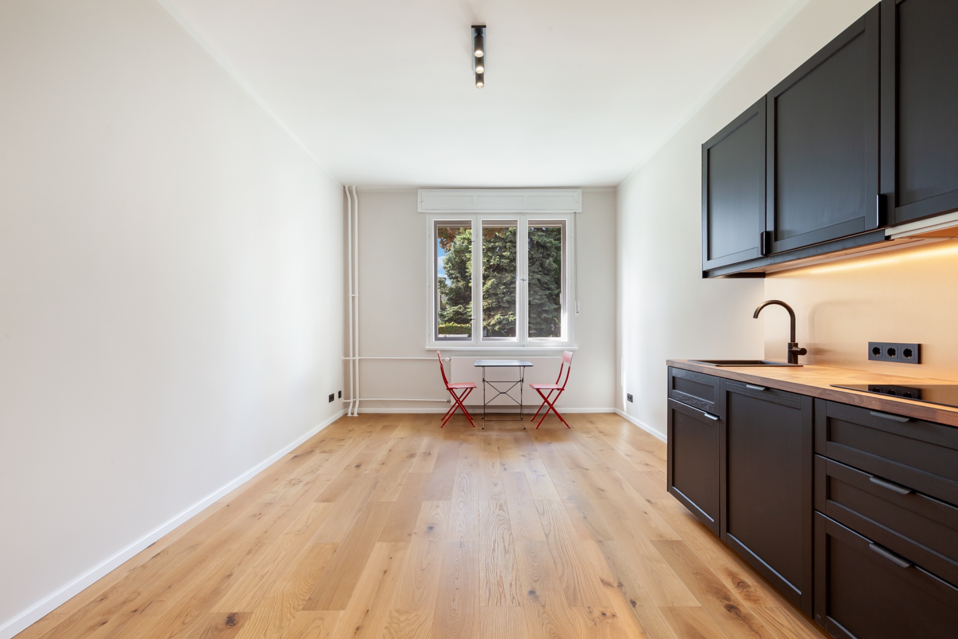 Heller Raum mit Holzboden, schwarzer Küchenzeile und kleinem Tisch mit zwei roten Stühlen am Fenster.
