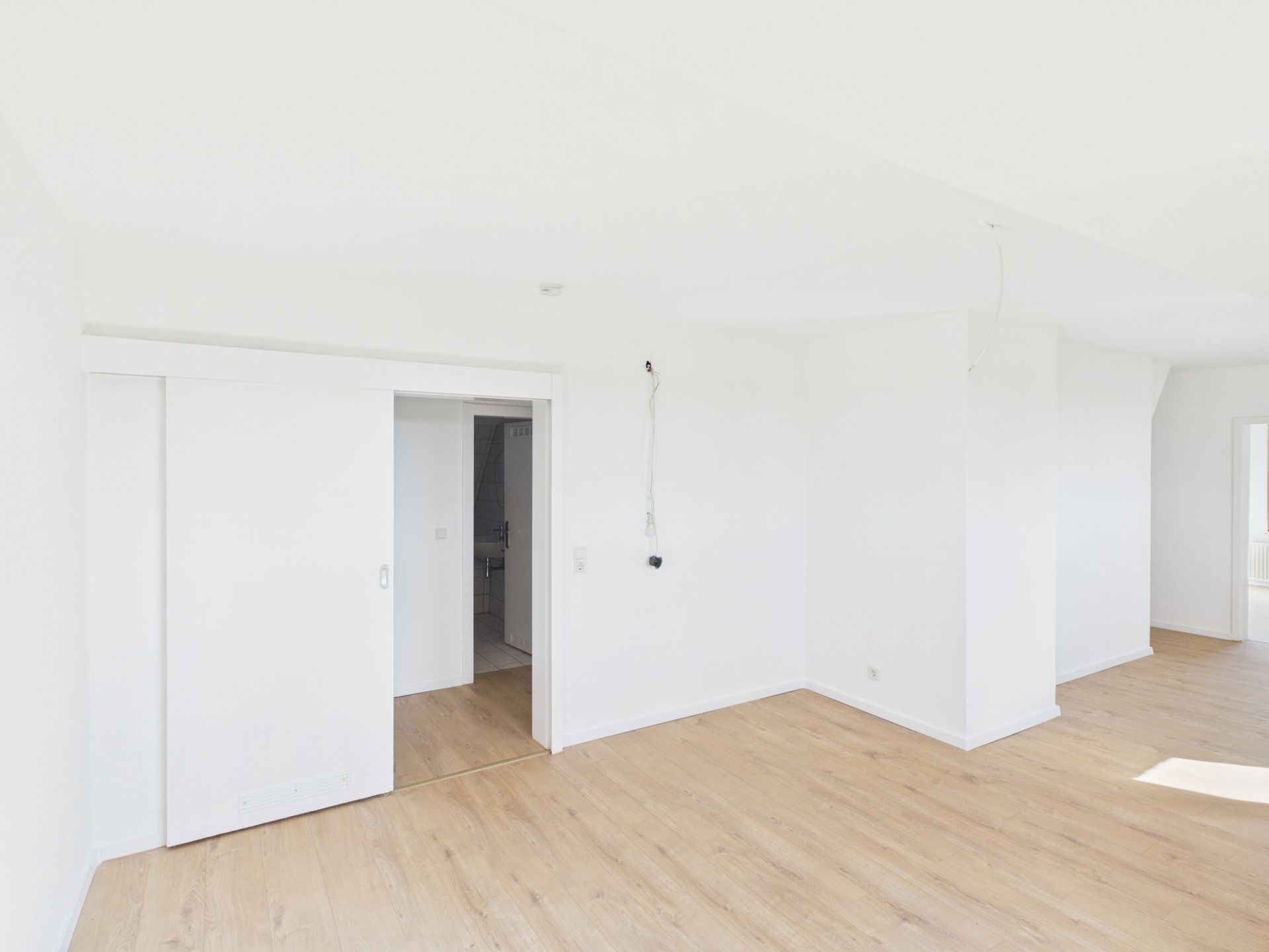 Helles, leeres Zimmer mit Holzfußboden und weißen Wänden, Schiebetür und viel Tageslicht.