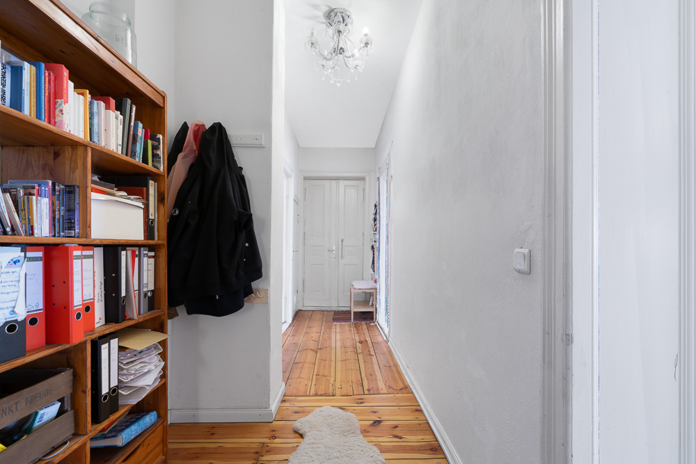 Heller Flur mit Holzregal voller Bücher und Aktenordner, Holzboden und Garderobe an der Wand.