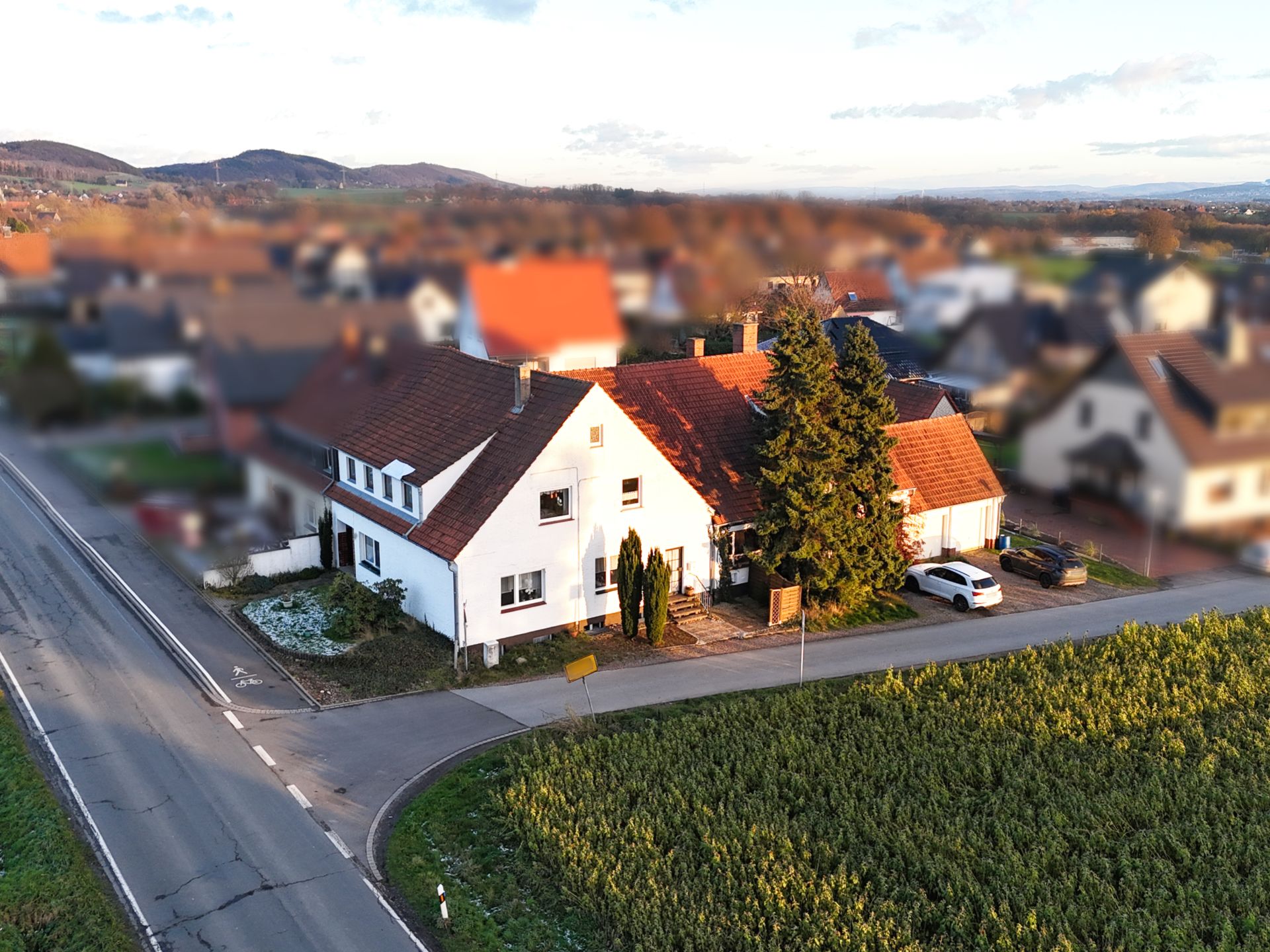 Weißes Wohnhaus mit rotem Dach an einer Straße, daneben ein Baum und ein geparktes Auto.