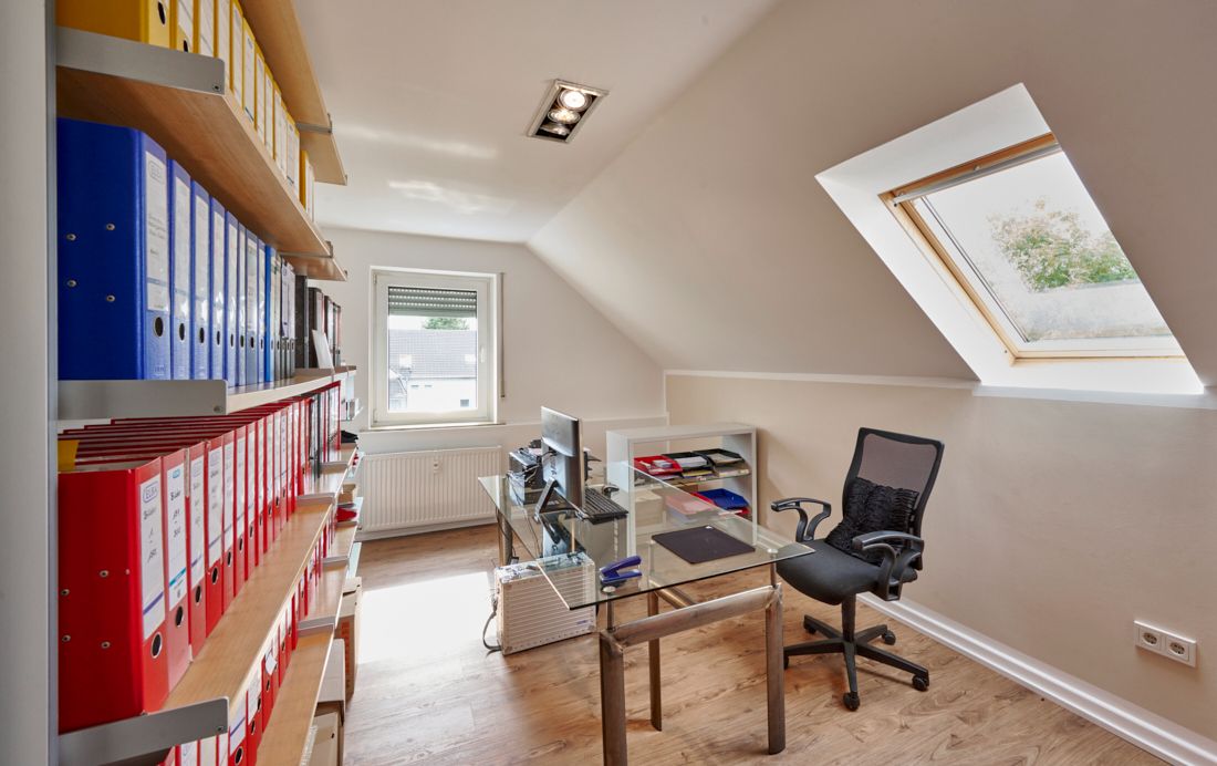 Helles Arbeitszimmer mit Schreibtisch, Bürostuhl, Computer und vielen Ordnern im Regal.