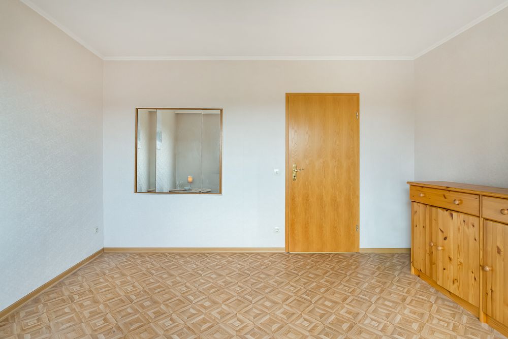 Leerer Raum mit Holztür, Fenster zum Nebenraum und Holzkommode auf Parkettboden.