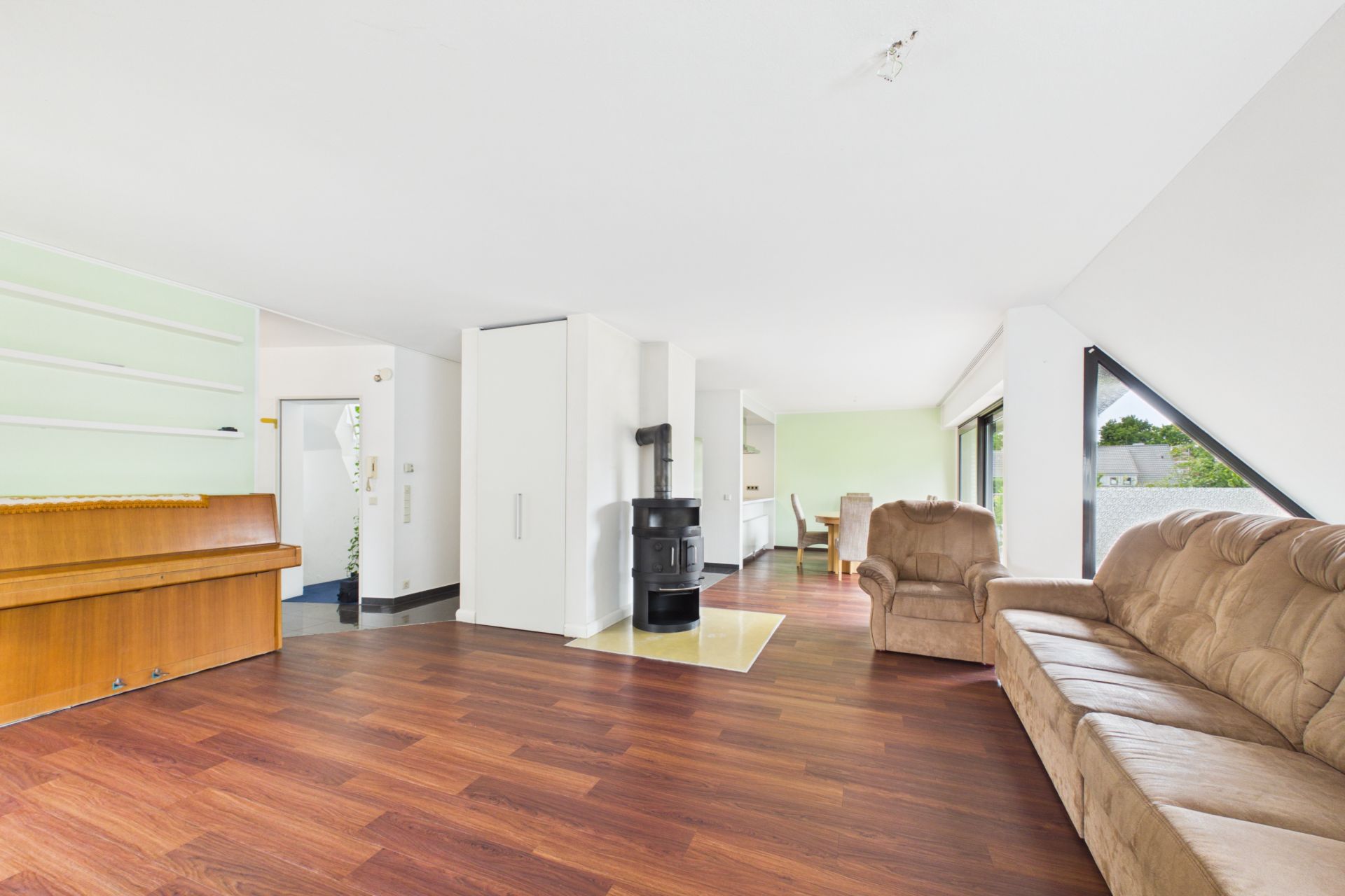 Helles Wohnzimmer mit braunem Sofa, Sessel, Kaminofen, Klavier und großen Fenstern.