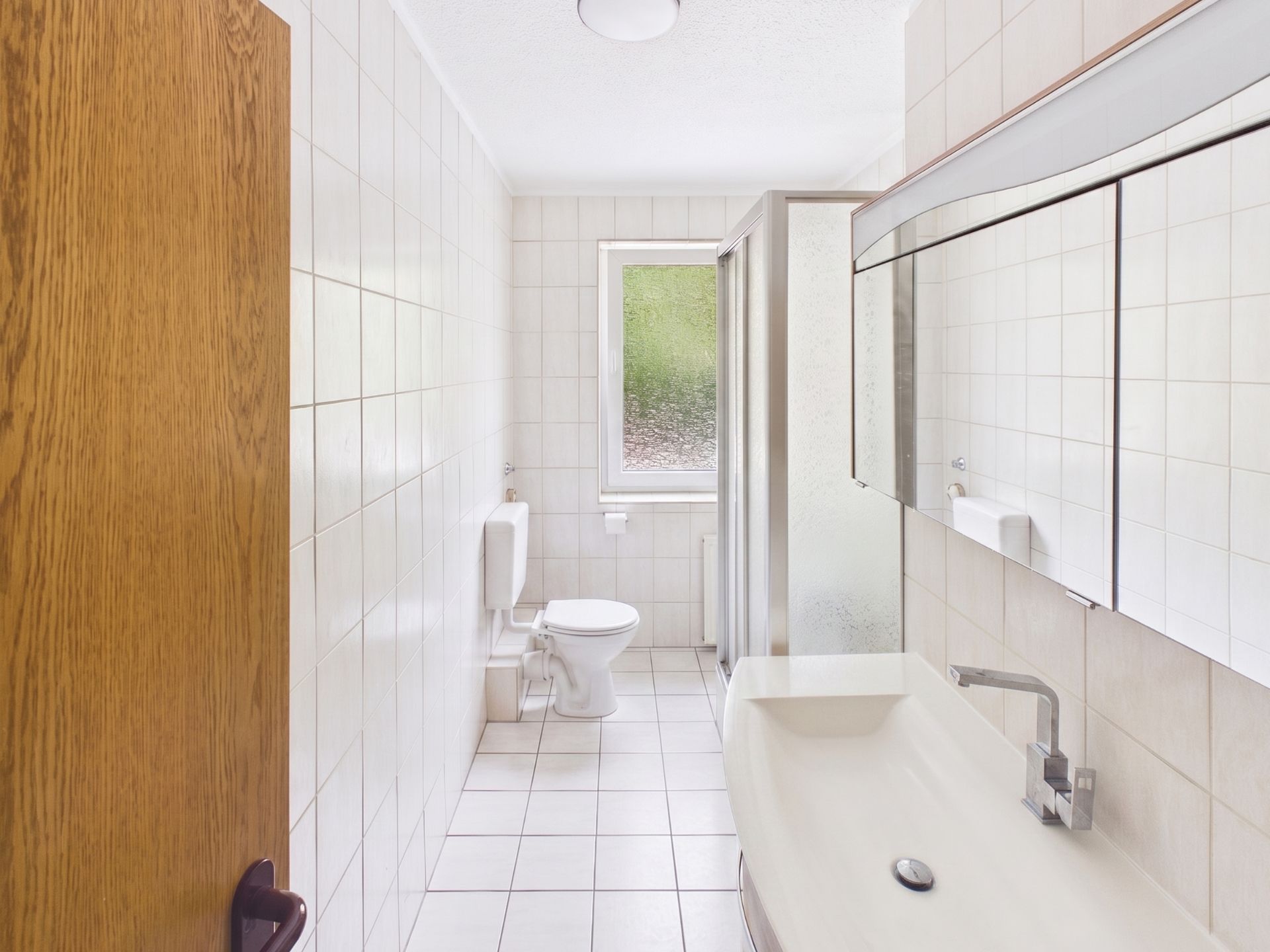Helles Badezimmer mit Dusche, Toilette, Waschbecken und großem Spiegel, gefliest und mit Fenster.