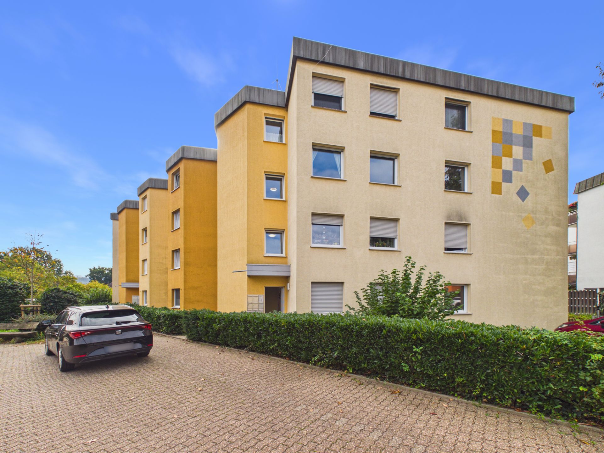 Mehrfamilienhaus mit gelber und beiger Fassade, Parkplatz und Auto im Vordergrund.