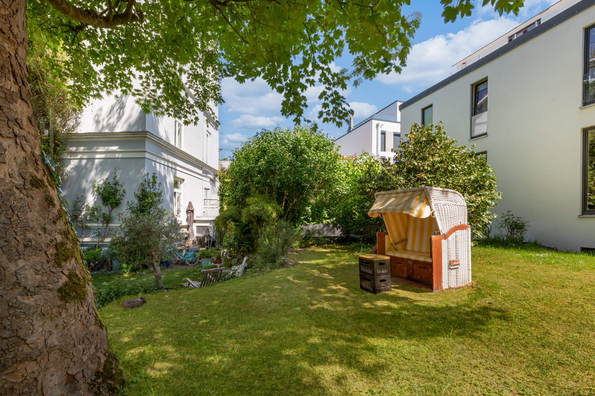 Grüner Garten mit Liegestuhl, Bäumen und angrenzenden Wohnhäusern an einem sonnigen Tag.