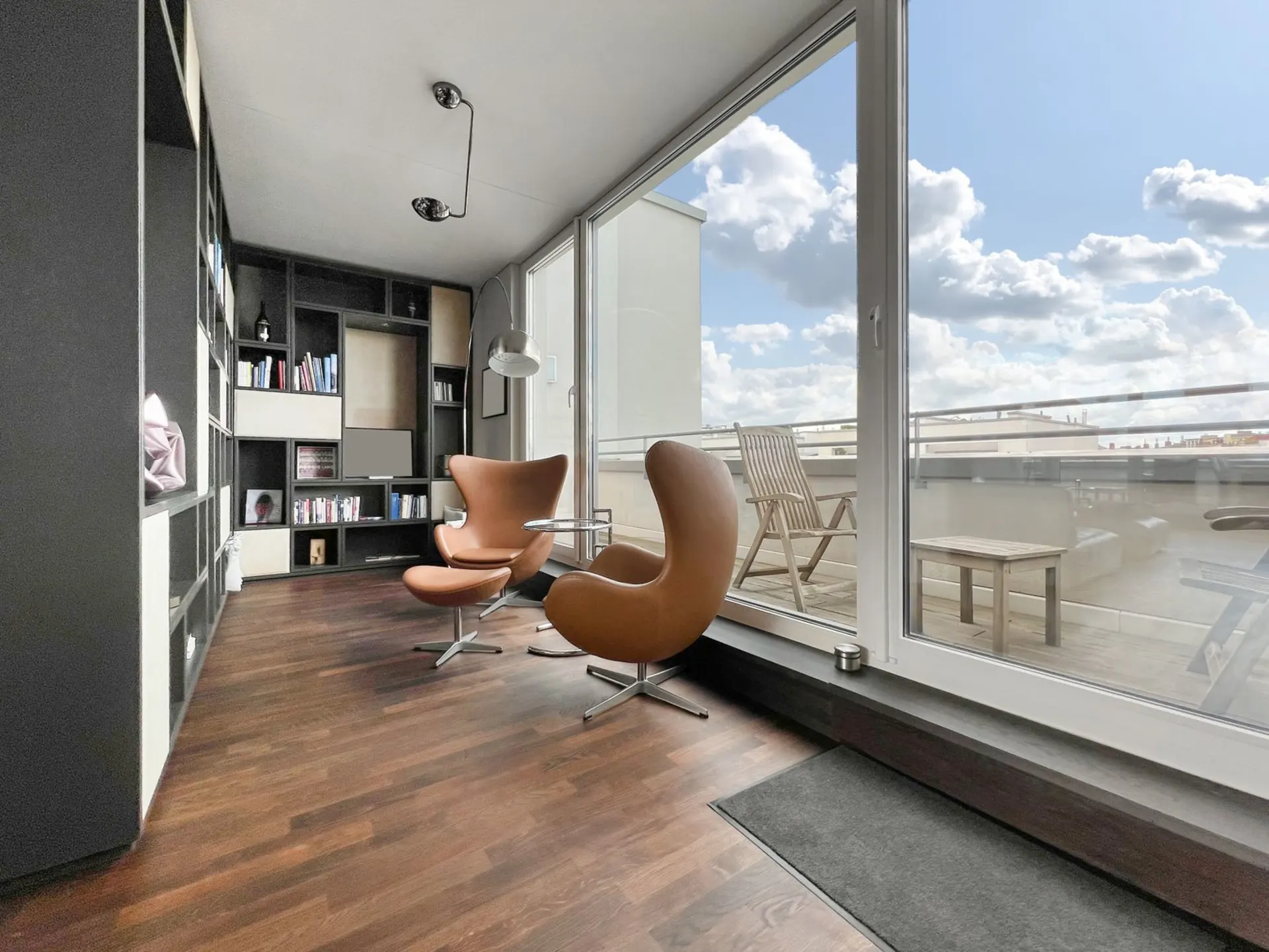 Wohnzimmer mit zwei braunen Sesseln, Bücherregal und großer Fensterfront zur Terrasse.