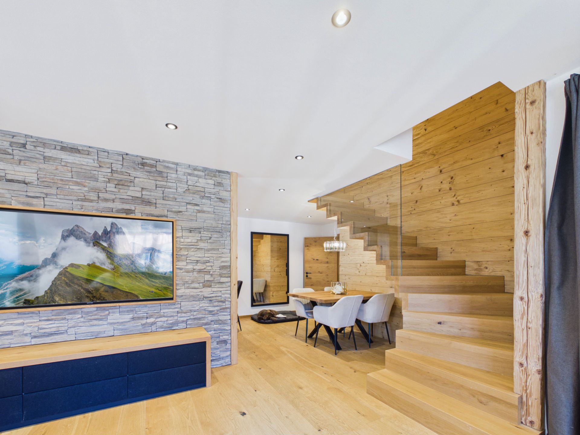 Modernes Wohnzimmer mit Holztreppe, Esstisch und Natursteinwand mit Landschaftsbild.