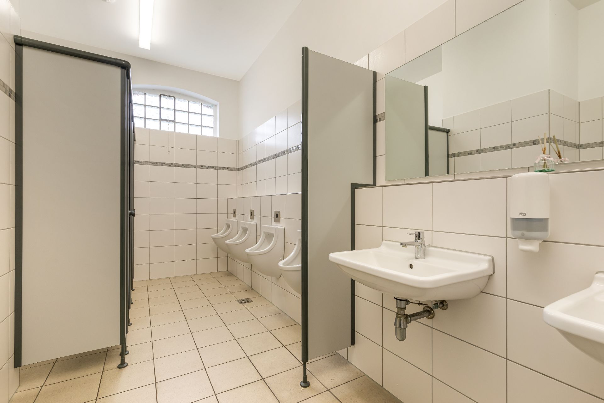 Heller, gefliester Waschraum mit Urinalen, Waschbecken und Spiegel an der Wand.