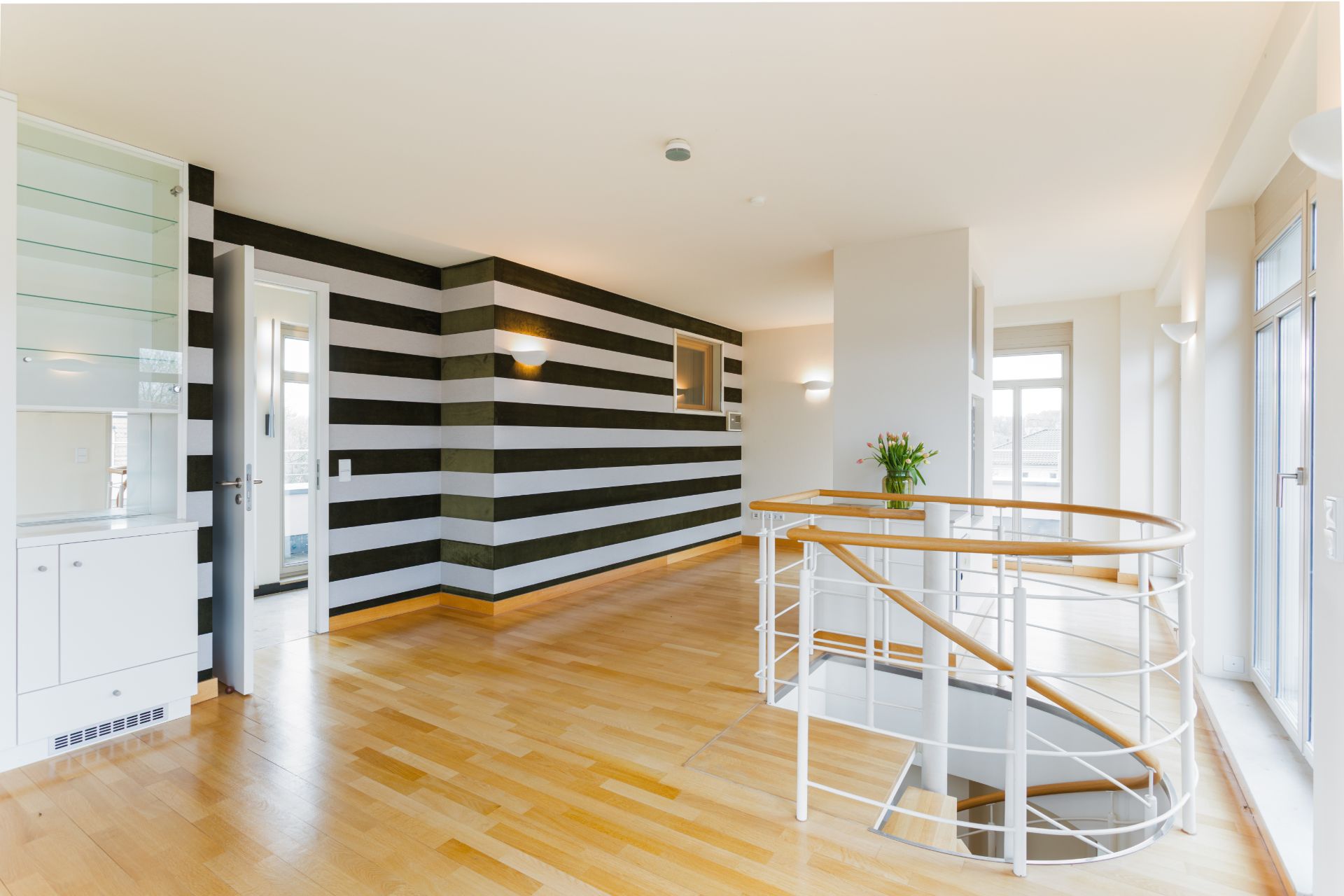 Heller Flur mit Holzfußboden, Treppengeländer und schwarz-weiß gestreifter Wand.