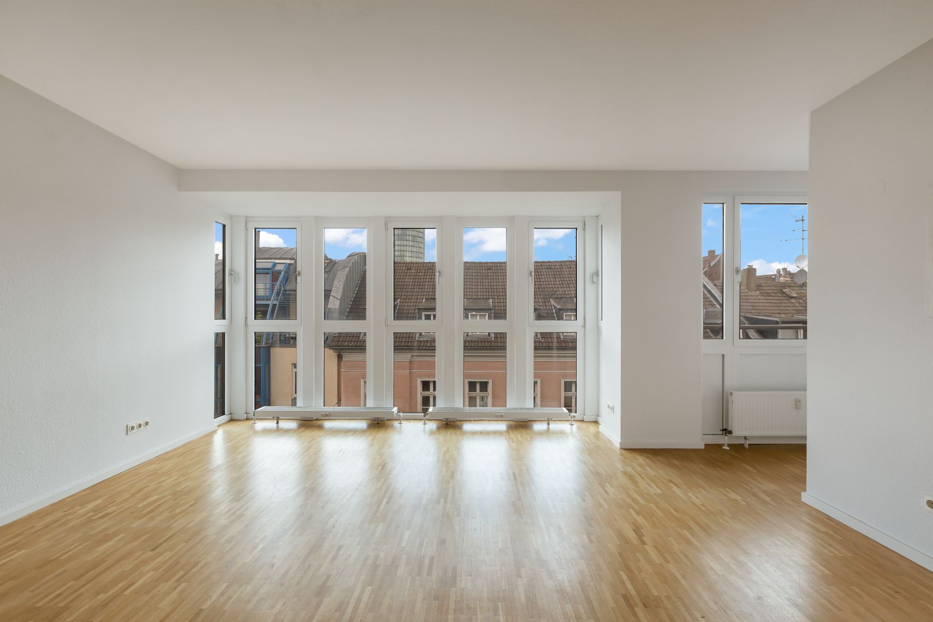 Helles, leeres Wohnzimmer mit Parkettboden und großen Fenstern, Blick auf Häuserdächer.