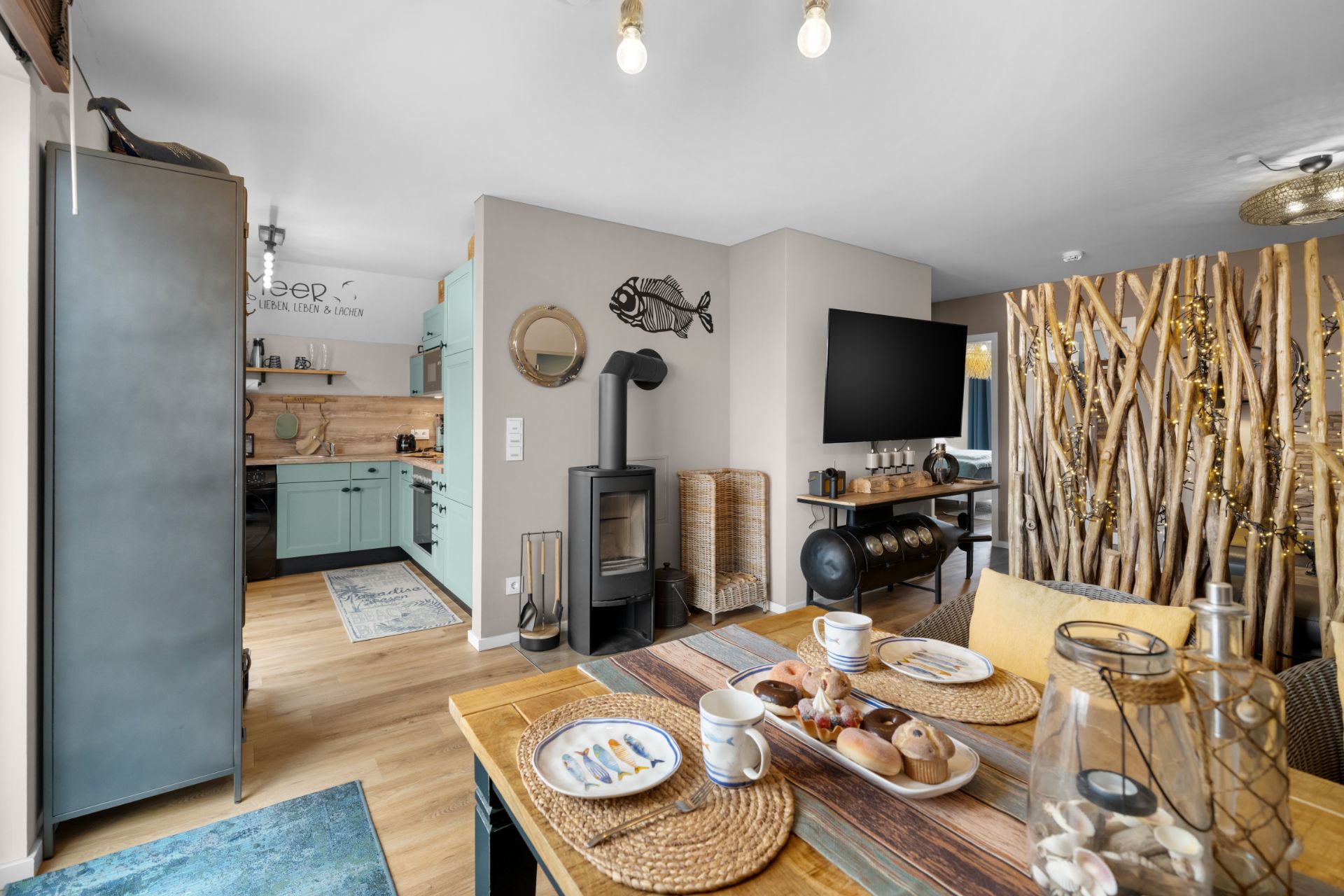 Gemütliches Wohnzimmer mit Esstisch, gedeckt für Frühstück, daneben eine offene Küche und ein Holzofen.