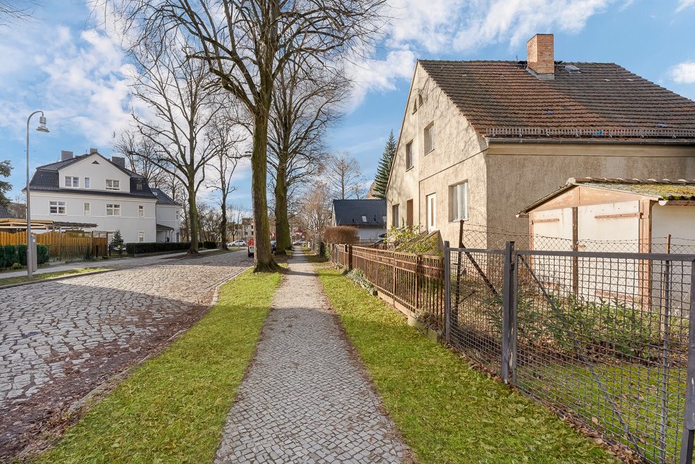 Gepflasterter Gehweg entlang eines Wohnhauses mit Gartenzaun, Bäumen und weiteren Häusern im Hintergrund.