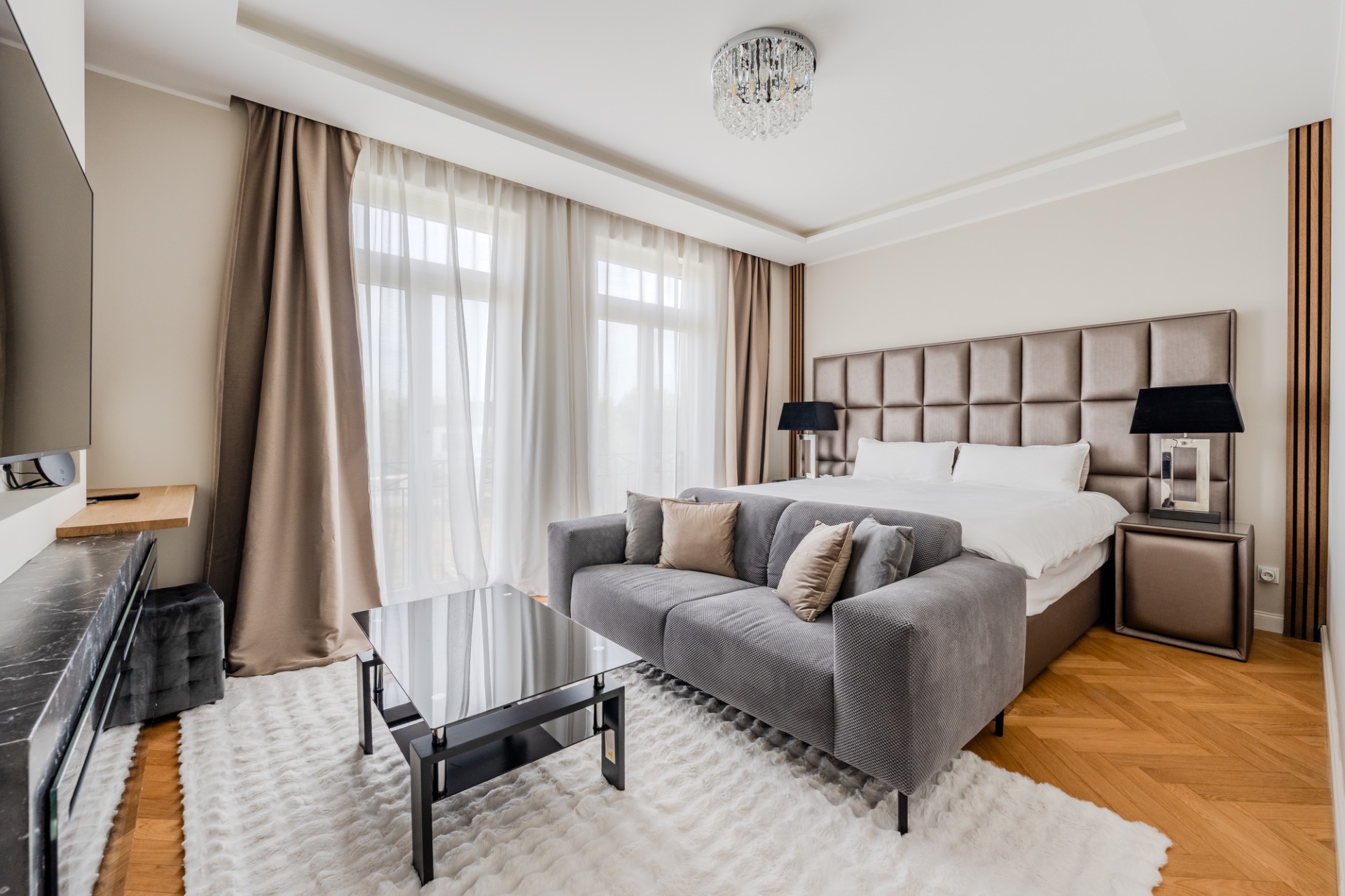 Modernes Schlafzimmer mit großem Bett, grauem Sofa, Couchtisch und großen Fenstern mit Vorhängen.