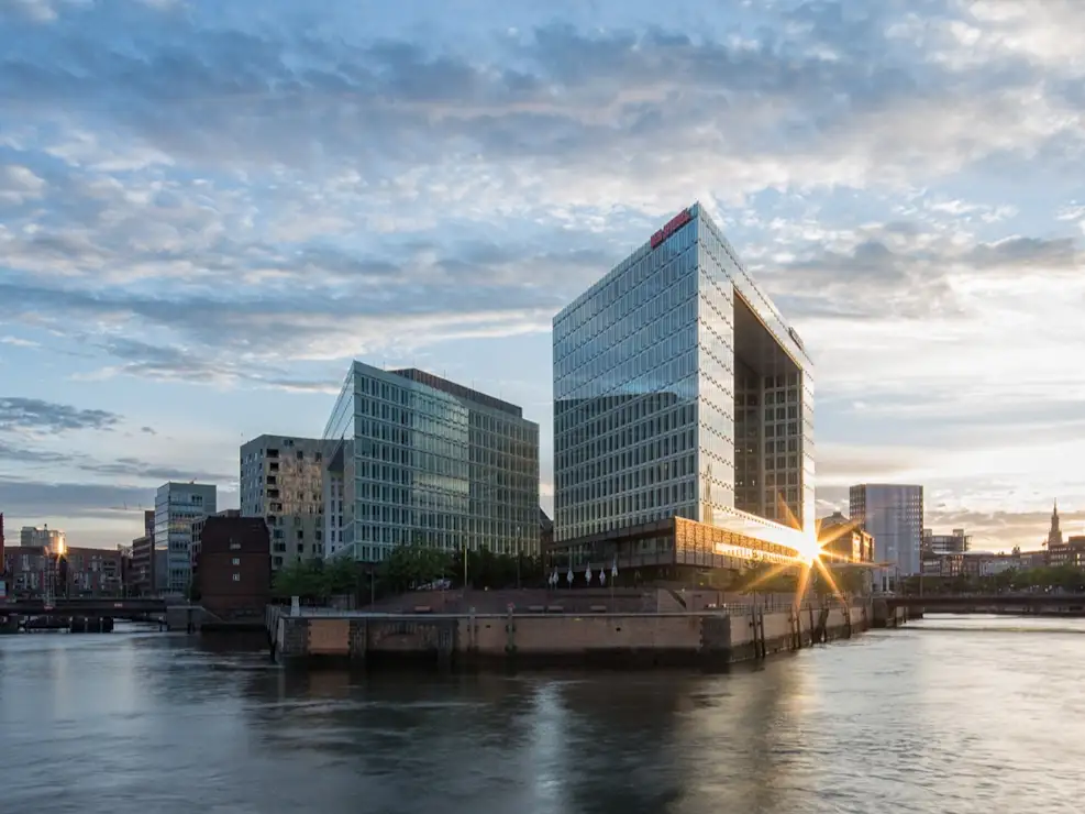 Moderne Glasgebäude am Wasser bei Sonnenuntergang, im Vordergrund eine Brücke.