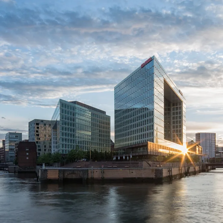 Moderne Glasgebäude am Wasser bei Sonnenuntergang, im Vordergrund eine Brücke.