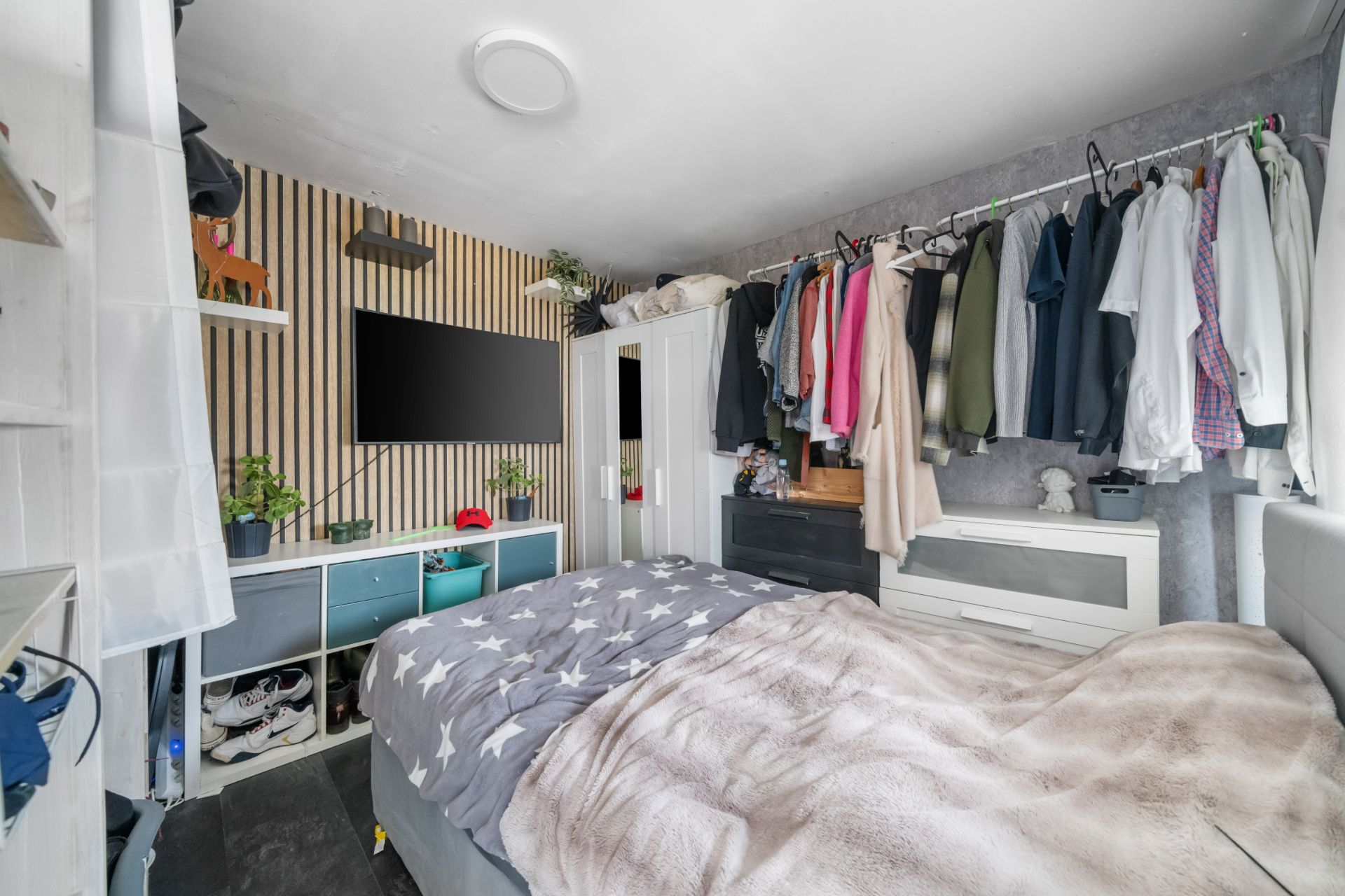 Schlafzimmer mit Doppelbett, Wandfernseher, offenen Regalen und Kleiderstange mit Kleidung.