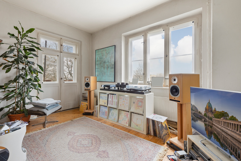 Helles Wohnzimmer mit großen Fenstern, Teppich, Pflanze, Schallplattenspieler und Lautsprechern.