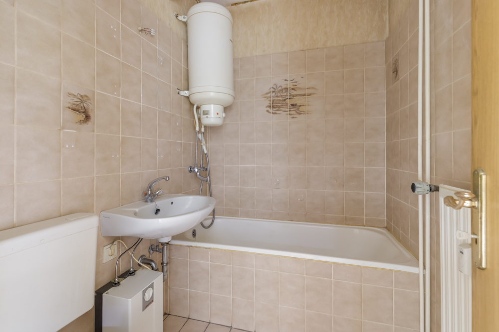 Kleines Badezimmer mit Badewanne, Waschbecken und Boiler an beige gefliester Wand.