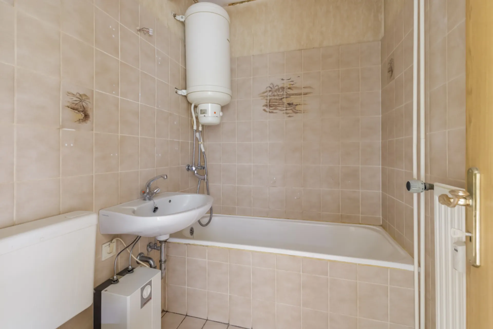 Kleines Badezimmer mit Badewanne, Waschbecken und Boiler an beige gefliester Wand.