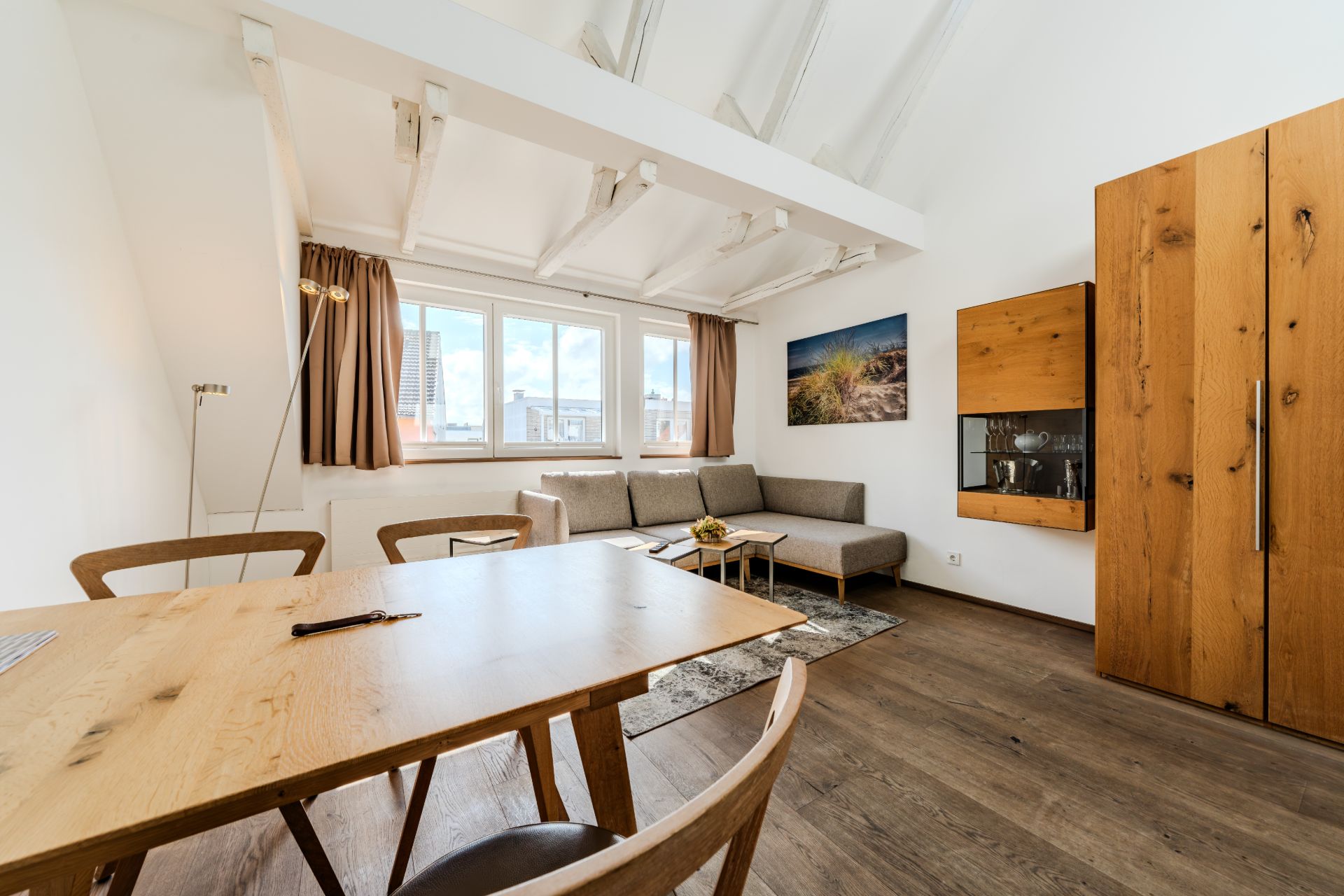 Helles Wohnzimmer mit Holztisch, Stühlen, Ecksofa, großem Fenster und Holzschrank.