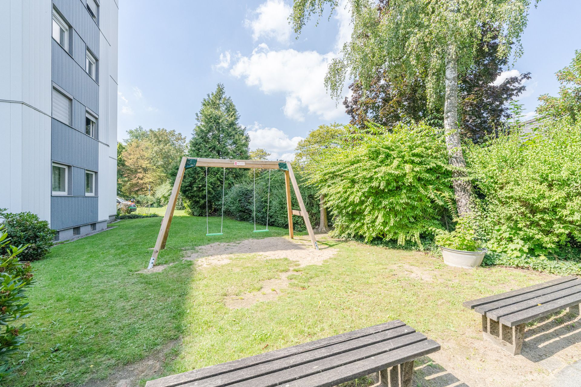 Gartenbereich mit zwei Schaukeln und Bänken neben einem Wohngebäude.