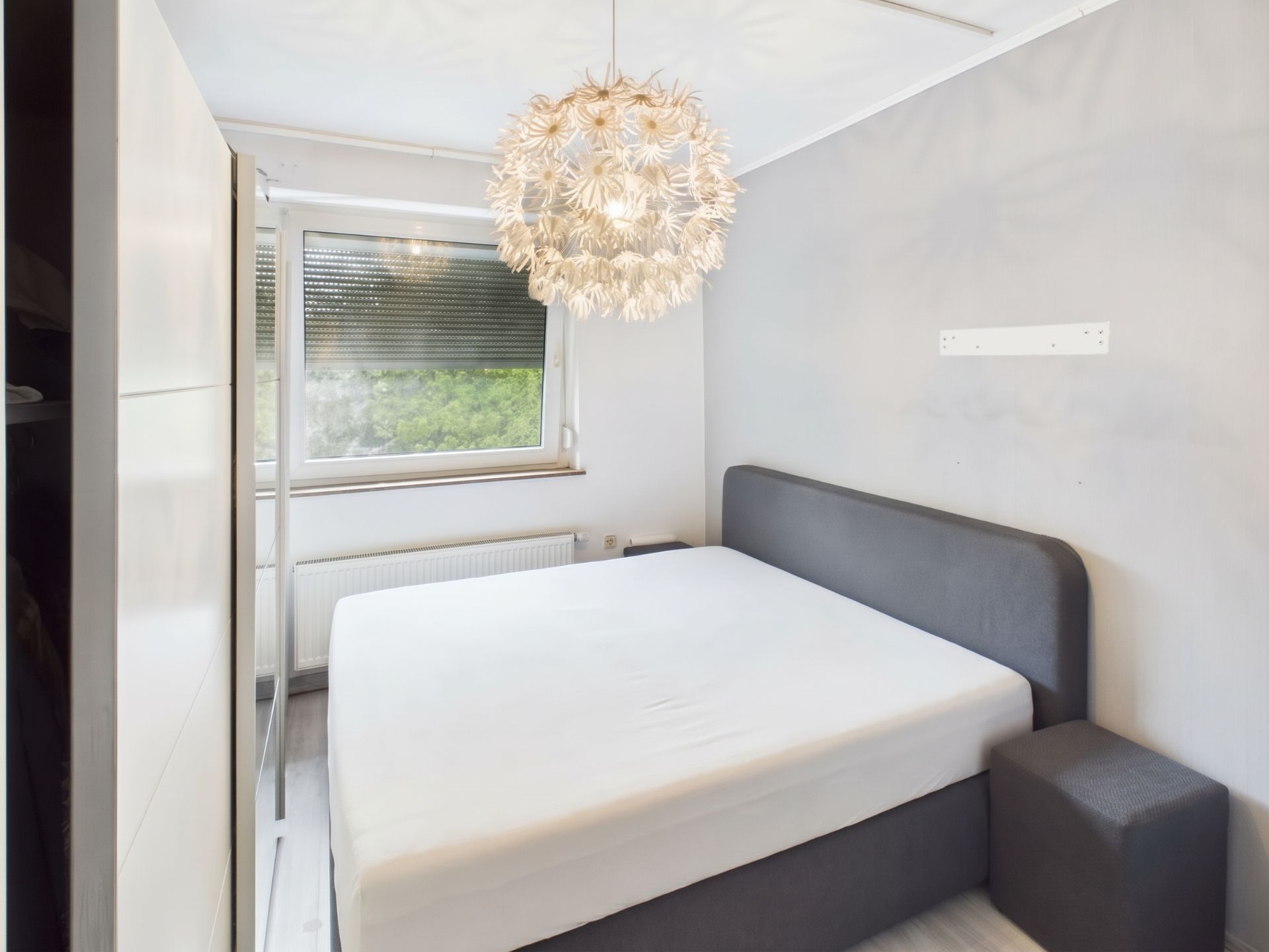 Schlafzimmer mit grauem Bett, großem Fenster und auffälliger Deckenleuchte.