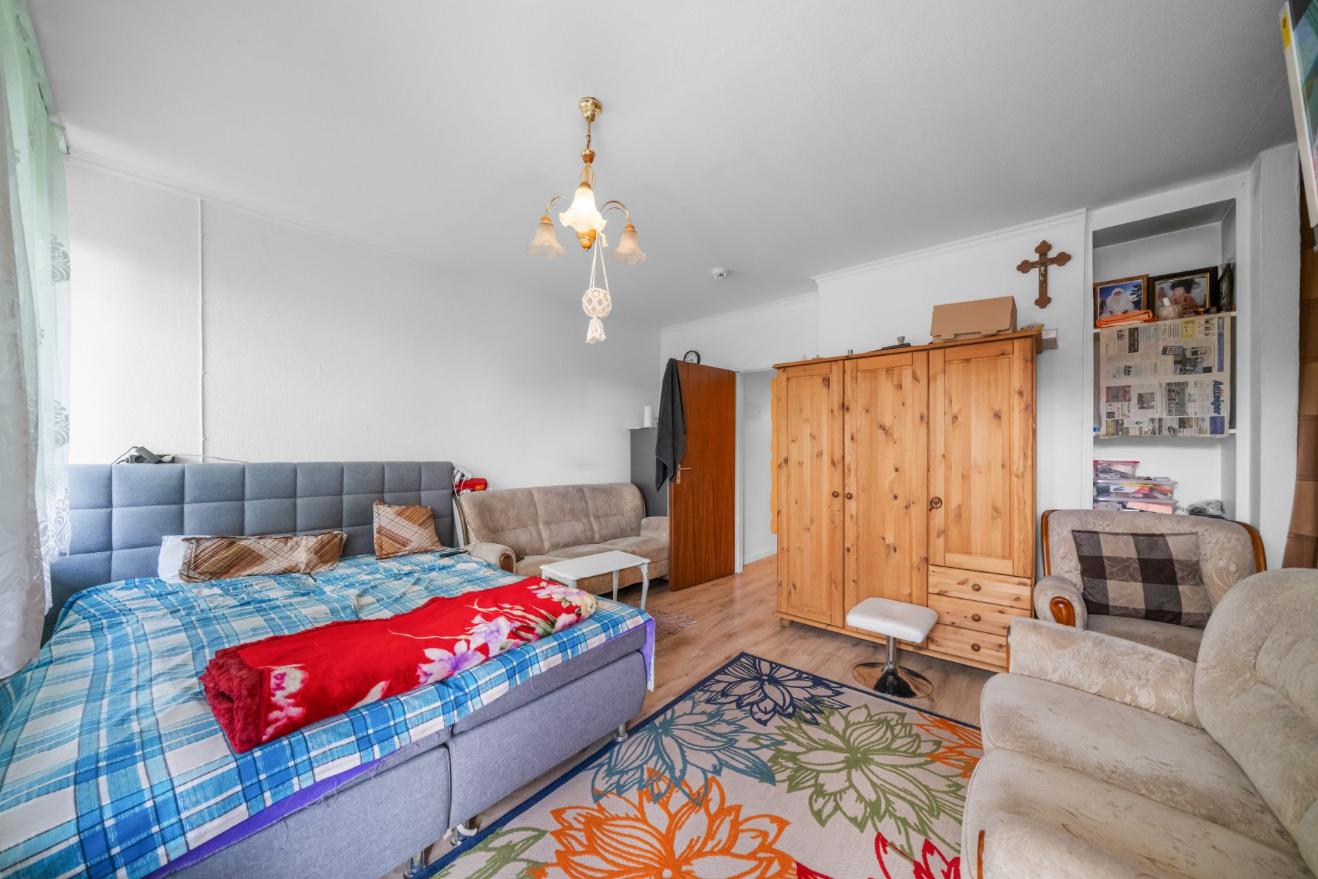 Wohnzimmer mit Bett, Sofa, Holzschrank, Regal und bunter Teppich mit Blumenmuster.